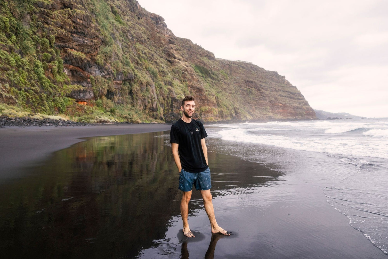 Lucas barefoot on a black sand beach below green cliffs