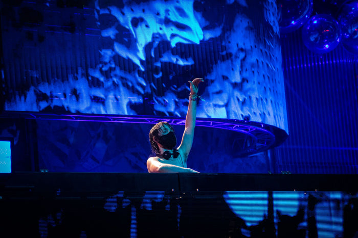 DJ with a mask raising his arm in front of a huge blue screen
