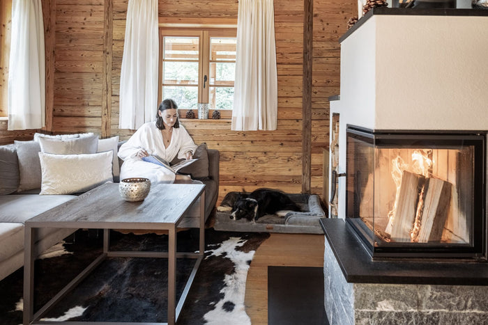 Guest in a bathrobe reading by the fireplace in a wooden chalet, dog resting beside her