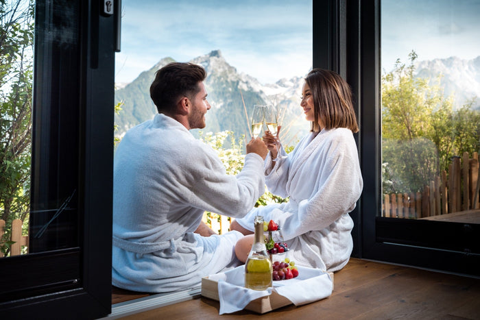Couple in bathrobes toasting with champagne at a large window with mountain view