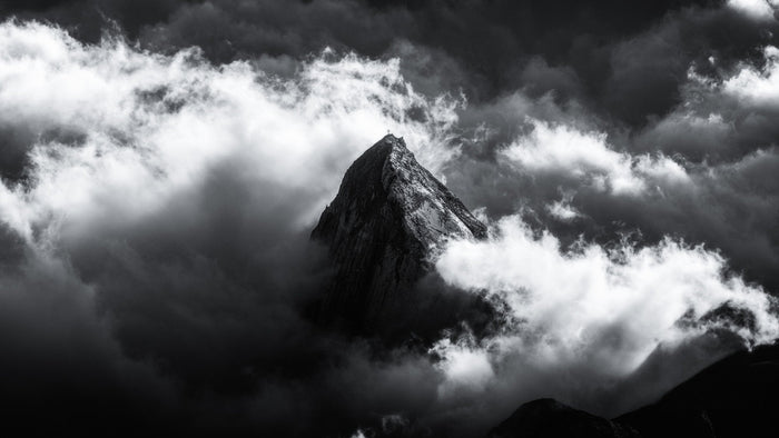 Black and white summit cross on a rock tower breaking through clouds