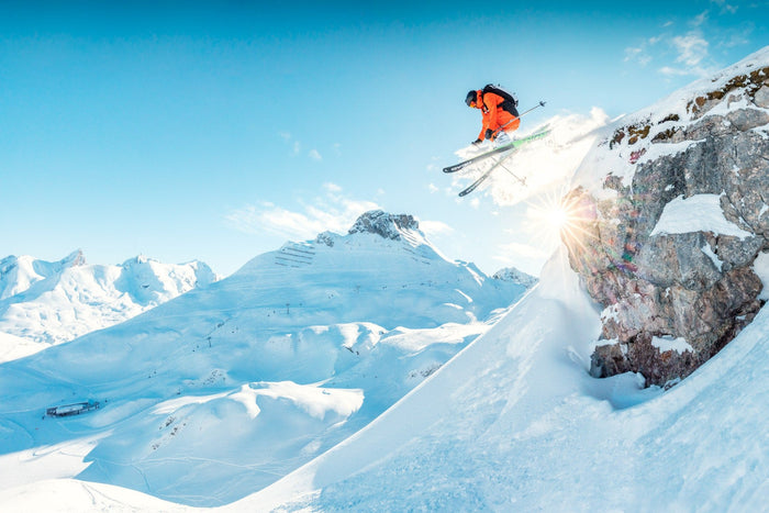 Skier in an orange jacket jumping off a rock into the sun