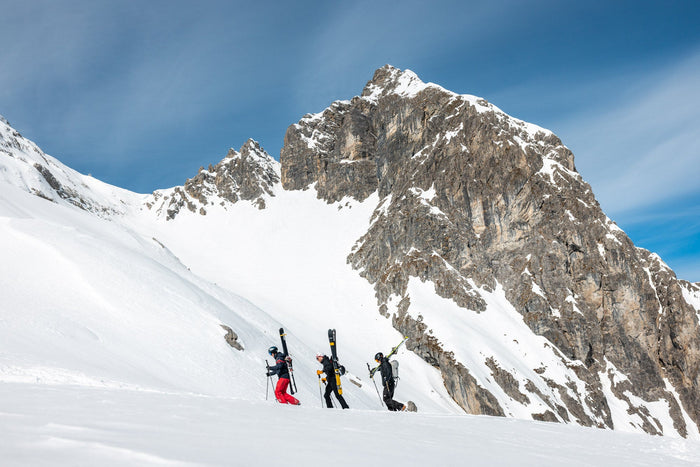 Three skiers bootpacking with skis on their backs below a rock peak