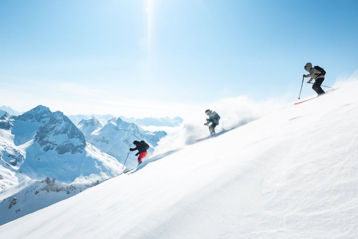 Three skiers descending a sunlit powder slope with mountains behind