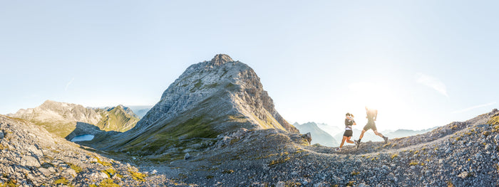 Panorama of two trail runners passing a peak and a mountain lake